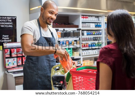 Smiling salesman putting vegetables in bag for customer after billing. Cashier black man at grocery store helping customer pack purchased products. Happy young man working in grocery shop.