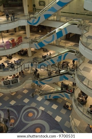 Shopping mall interior with escalators and meeting area (lobby). People faces, logos and information tables  blurred. Wide 5 floors view.