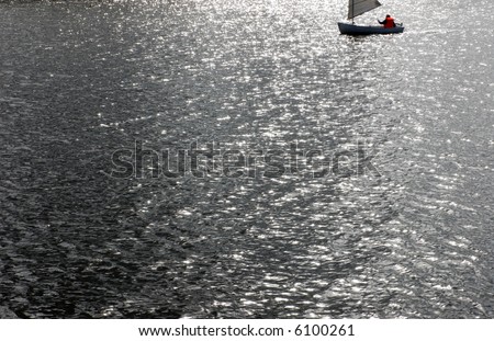 Lone sailor in a small sailboat sails across calm light dappled waters