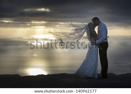 Bride and groom holding hands look at each other\'s eyes at sunset in the Canary Islands, the veil fluttering in the wind.