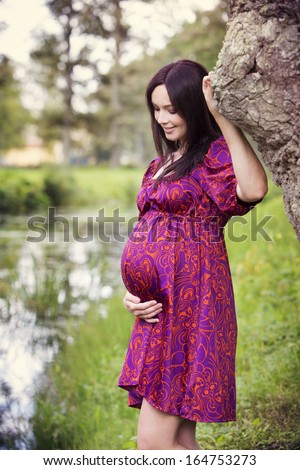 Happy pregnant woman standing near the pond and embracing her belly.