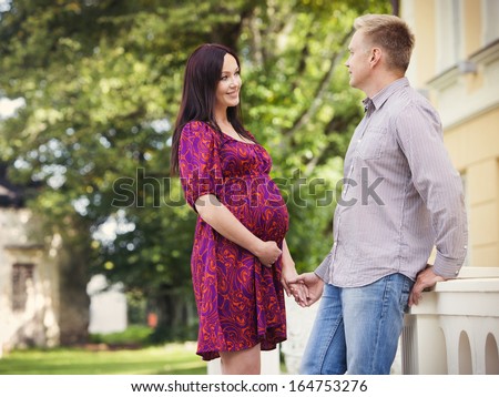 Happy pregnant woman standing with her husband holding hands outdoors in the Summer.