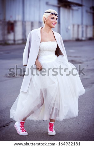 Beautiful bride with a fashionable eye-catching make-up standing on the street wearing a wedding dress and casual shoes.
