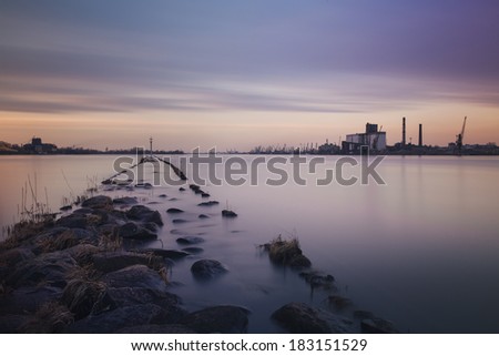 Evening landscape with beautiful colors in the sky, a peaceful river and the mole distant shore industrial landscape with chimneys and containers.