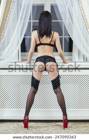 Sexy young woman from back with long brown hair in black lingerie leaning against the window sill and look through the window.