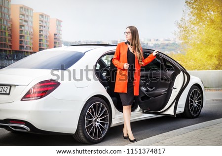 Attractive businesswoman with glasses and red coat gets out of the white Mercedes S-Class car, modern city background with rear view of the car