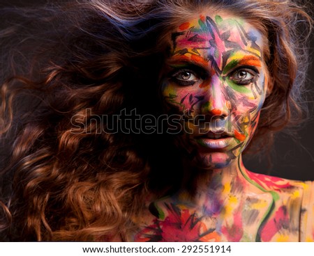 Young beautiful woman with flying hair bright glowing eyes and tribal colorful face art painted face on dark grey background