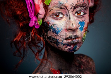young woman with glamorous bizarre face art red curly hair with clips and glowing brown eyes on dark blue background in studio