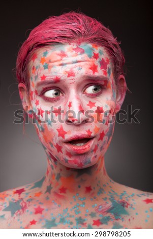 Emotional portrait of a surprised or annoyed woman looking like a fabulous creature with stars on the face and painted hair in pink in studion on grey background