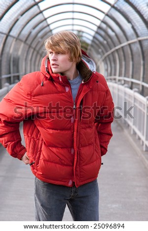 A male model with his hands at his sides on a covered walking bridge wearing a bright red jacket.