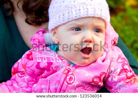 A young infant child yells with her mouth open while her mom holds her outdoors.