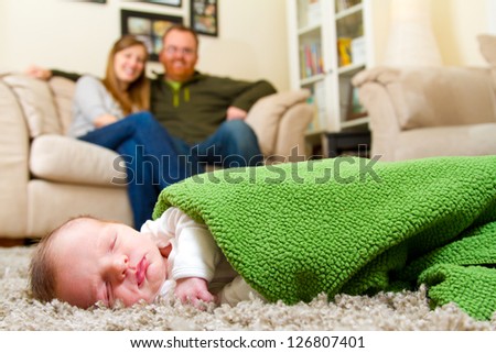 A newborn baby boy sleeps on the floor with his parents behind him slightly out of focus. The three people are in the living room of the mother and father\'s house.
