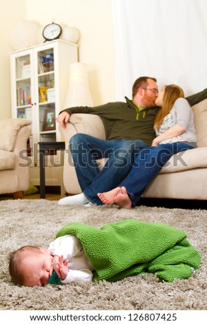A newborn baby boy sleeps on the floor with his parents behind him slightly out of focus. The three people are in the living room of the mother and father\'s house.