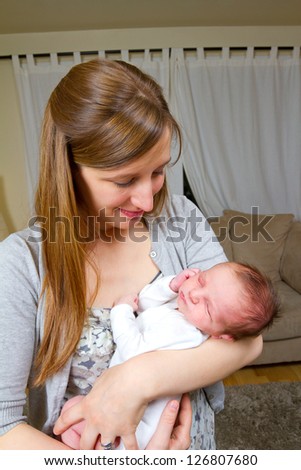 A mother holds her newborn baby in her arms while looking down at him with a happy look. The baby boy is in comfort and trusts his mom fully. He is sleeping peaceful.y.