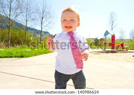 A baby boy plays at the park wearing his Sunday\'s best clothes including a tie around his neck.