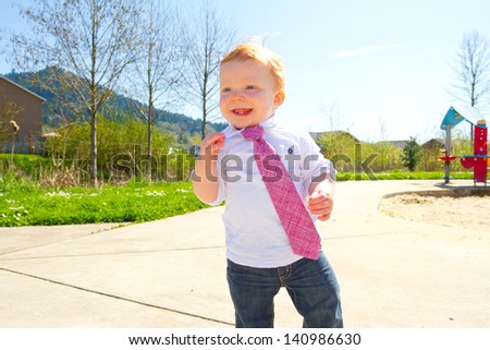 A baby boy plays at the park wearing his Sunday\'s best clothes including a tie around his neck.