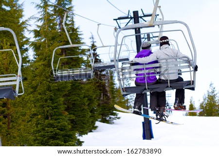 A couple of people ride the ski chair lift up the mountain together while sitting closely to each other having a fun time during a day of snowboarding in oregon.