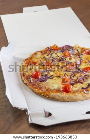 Delivered pizza with tomatoes and onions in a open pizza box on a brown wooden table
