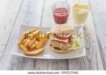 homemade burger and french fries on a white plate with ketchup and mayonnaise in glasses on a wooden table