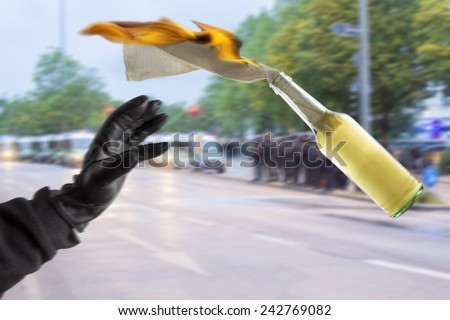 Arm in black sleeve with black leather glove thwowing a Molotov Cocktail with burning fuse, with police forces in the blurred background