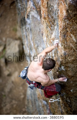A male climber, viewed from above, climbs a very high and steep crag. Shallow depth of field is used to isolate the climber with focus on the hands and head