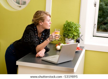 A woman using a laptop in the kitchen - looking out the window as though she is thinking about someone or something.