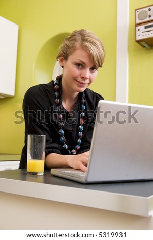 A young woman uses the computer in the kitchen while enjoying a glass of juice. The model is looking at the viewer and smiling.