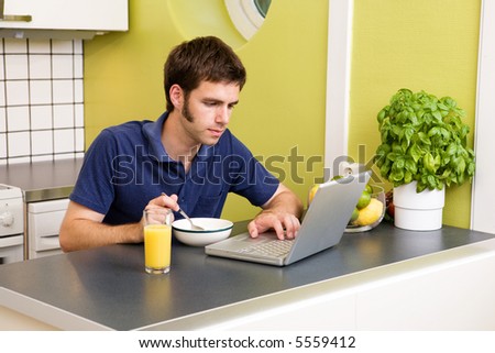 A young male works on the computer in the kicthen while eating breakfast