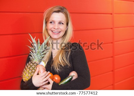 Smiling woman leaning against red wall while holing fruits and vegetables