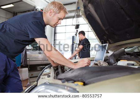 Mechanic using laptop while working on car with people in background