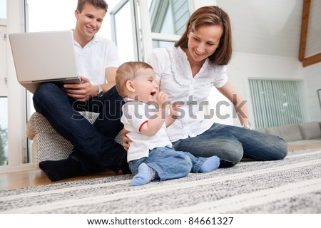 Smiling mother playing with cute child with man using laptop in background