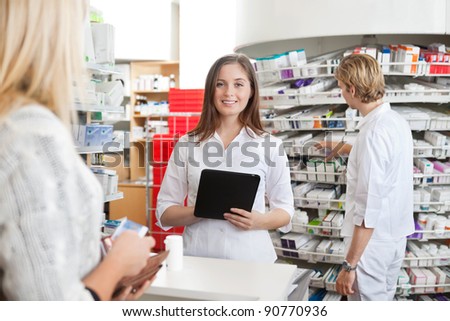 Portrait of female pharmacist holding tablet PC while attending customer on the counter