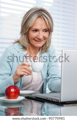 Smiling mature woman working on laptop while having cup of tea