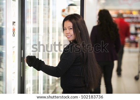 Young woman smiling while opening refrigerator in supermarket with people in the background