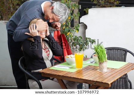 Middle aged man covering woman\'s eyes on breakfast table for surprise gift