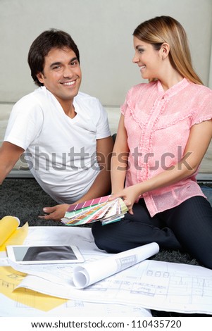 Portrait of happy young man with woman holding color swatches while sitting on rug with tablet PC and blueprints