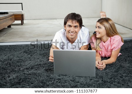 Portrait of happy young man lying on rug with laptop while woman looking at him