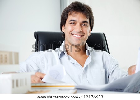 Portrait of happy young male architect with blueprint sitting in office chair