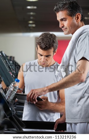 Male instructor making notes on clipboard while standing besides man on treadmill in health club