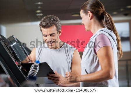 Happy male instructor holding clipboard while standing besides client exercising on treadmill in health club