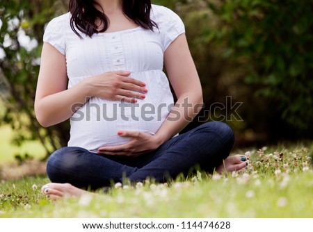 Faceless pregnant woman in third trimester sitting in park holding belly