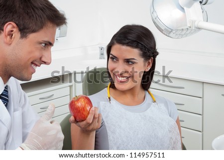 Happy male dentist showing thumbs up sign to a female patient holding an apple in clinic