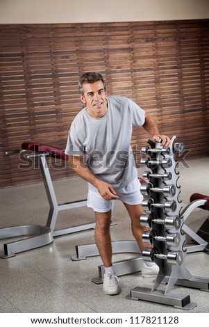 Portrait of young man standing by dumbbells in rack at health club