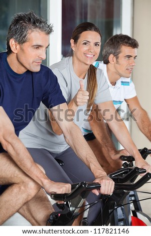 Portrait of happy mature woman showing thumbs up sign while exercising with friends on spinning bike in health club