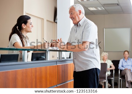 Senior man communicating with female receptionist while women sitting in background Senior man communicating with female receptionist while women sitting in background