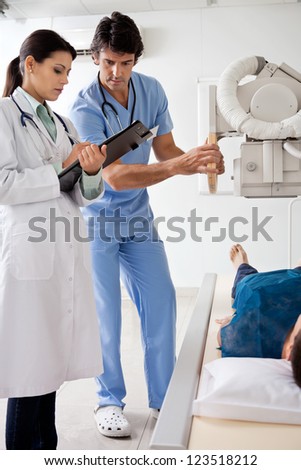 Female radiologist with technician looking at clipboard while setting up the machine to x-ray male patient