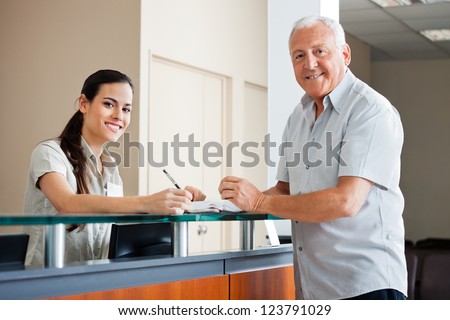 Portrait of senior man standing at hospital reception while female making an entry in book