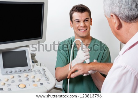 Young male radiologic technician smiling while scanning male patient\'s hand at clinic
