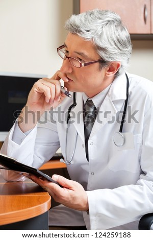 Thoughtful mature male doctor with clipboard sitting at desk in clinic