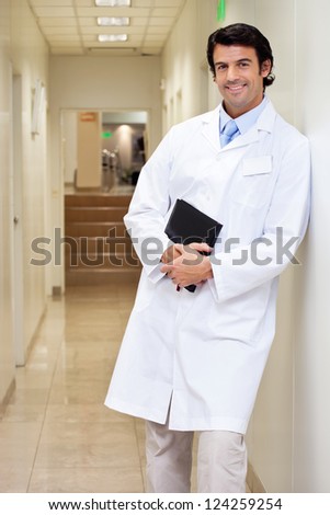 Portrait of a happy mixed race male doctor holding book while standing in hospital passageway